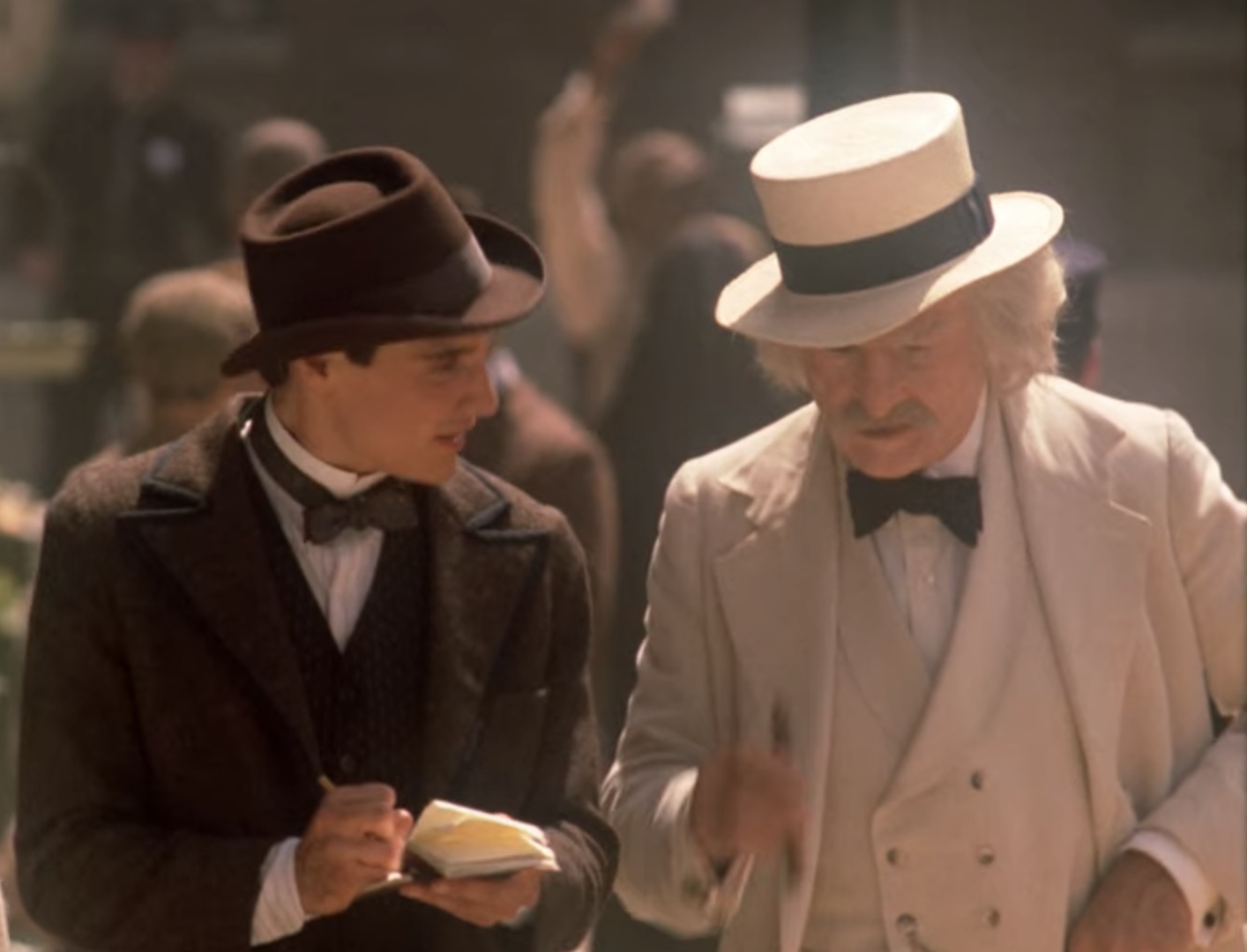 Still image from Star Trek TNG episode Time's Arrow, Part II showing a close-up of Samuel Clemens (aka Mark Twain) standing in the street, talking to a reporter holding a notepad sometime in the 1890s. Clemens is wearing a white three-piece with a white panama hat and holding a cigar. The reporter is wearing a dark brown suit with a homburg.